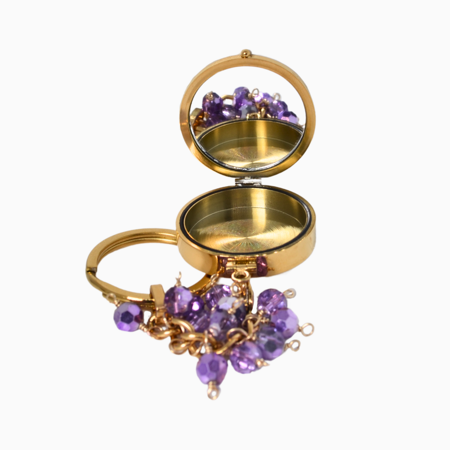 The Amethyst Pocket Locket Keychain opened to reveal its built-in mirror and hidden compartment. The gold stainless steel locket shines alongside its vibrant purple glass crystal, making it both stylish and functional for carrying small essentials.
