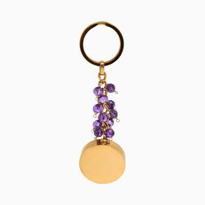 A gold stainless steel Amethyst Pocket Locket Keychain with a vibrant purple glass crystal, hanging elegantly. The locket is open to reveal a built-in mirror and a versatile compartment, perfect for storing small essentials. A thoughtful gift for February birthdays or anyone drawn to Amethyst’s calming energy.